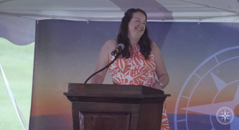 A woman with long brown hair is standing behind a wooden podium, smiling and looking to her right. She is wearing a white dress with an orange floral pattern. Behind her is a backdrop featuring a compass rose and a sunset scene. The setting appears to be outdoors, possibly under a tent or canopy.
